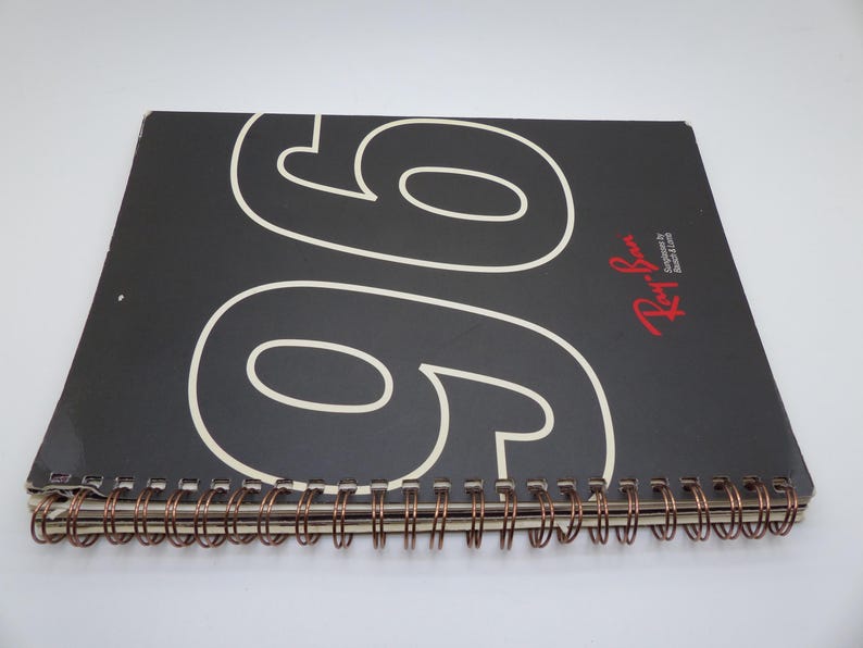 Puede incluir: Un cuaderno negro con espiral y el n&uacute;mero "96" en blanco en la portada. El logotipo de Ray-Ban es rojo. La encuadernaci&oacute;n es de color cobre.