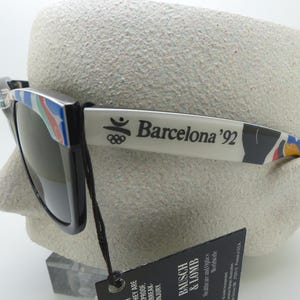 May include: Black sunglasses with the text "Barcelona '92" on the arm. The sunglasses have a colorful design on the frame and arm. The sunglasses are displayed on a mannequin head.