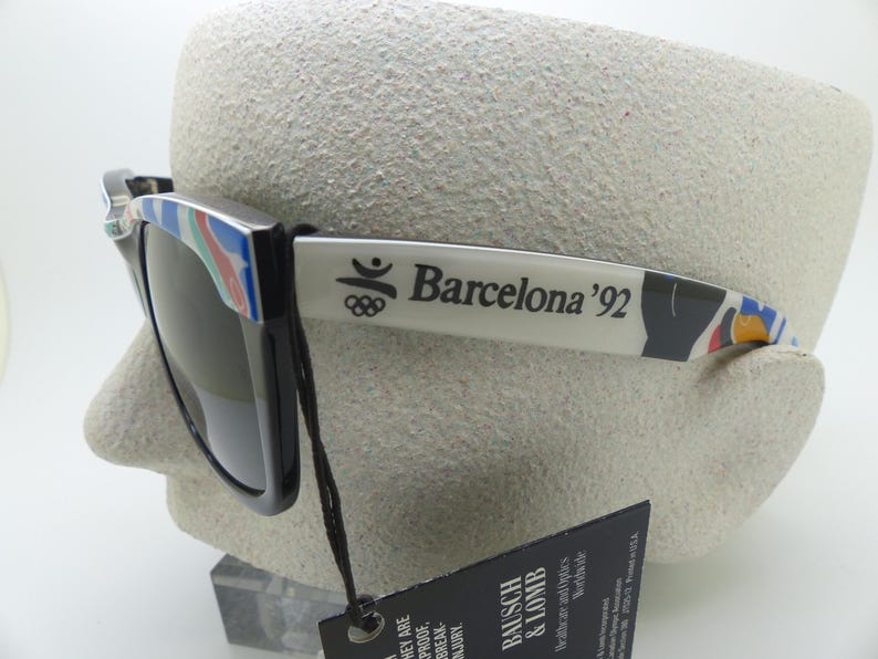 May include: Black sunglasses with the text "Barcelona '92" on the arm. The sunglasses have a colorful design on the frame and arm. The sunglasses are displayed on a mannequin head.