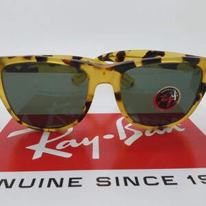 May include: Tortoiseshell-patterned sunglasses with dark lenses. The sunglasses are displayed on a red and white box with the Ray-Ban logo and the text "GENUINE SINCE 1937". A small sticker is on the right lens.