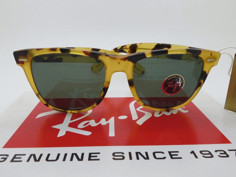 May include: Tortoiseshell-patterned sunglasses with dark lenses. The sunglasses are displayed on a red and white box with the Ray-Ban logo and the text "GENUINE SINCE 1937". A small sticker is on the right lens.