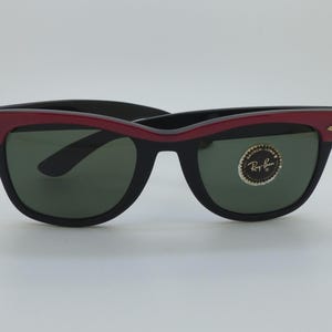 Puede incluir: Gafas de sol negras con montura superior roja y lentes verdes. Las gafas de sol tienen un dise&ntilde;o cl&aacute;sico con un peque&ntilde;o acento dorado en las patillas. Una pegatina circular est&aacute; en la lente derecha.