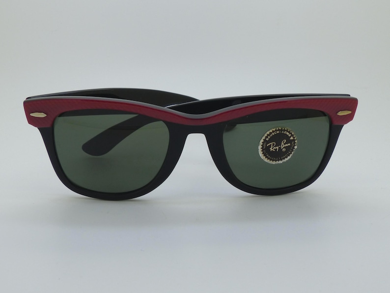 Puede incluir: Gafas de sol negras con montura superior roja y lentes verdes. Las gafas de sol tienen un dise&ntilde;o cl&aacute;sico con un peque&ntilde;o acento dorado en las patillas. Una pegatina circular est&aacute; en la lente derecha.