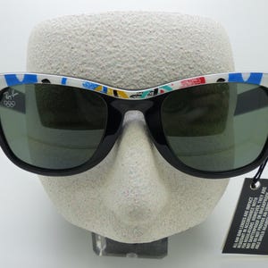 May include: Black Ray-Ban sunglasses with a colorful patterned design on the top of the frames. The sunglasses have green lenses and a tag from Bausch & Lomb. The Ray-Ban logo and Olympic rings are visible on the frame.