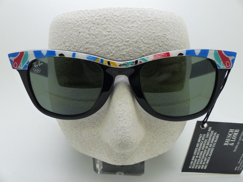 May include: Black Ray-Ban sunglasses with a colorful patterned design on the top of the frames. The sunglasses have green lenses and a tag from Bausch & Lomb. The Ray-Ban logo and Olympic rings are visible on the frame.