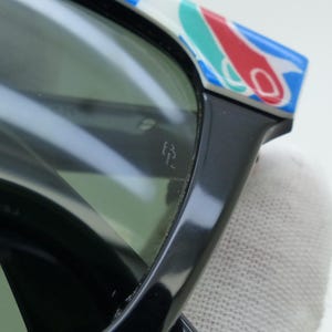 May include: Close-up of black sunglasses with green lenses. The top of the frame features a colorful pattern of red, blue, and white shapes. The glasses have a small logo etched on the lens.