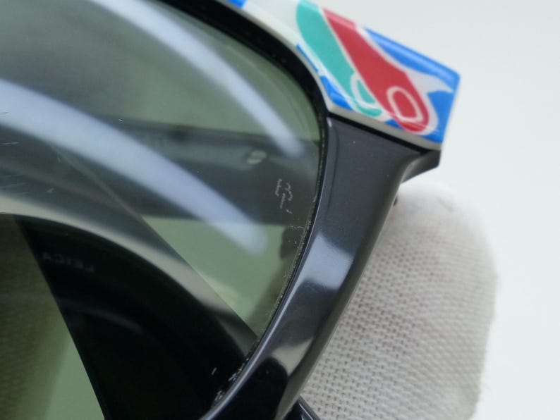 May include: Close-up of black sunglasses with green lenses. The top of the frame features a colorful pattern of red, blue, and white shapes. The glasses have a small logo etched on the lens.