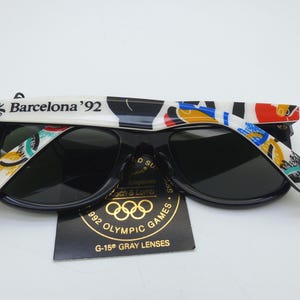 May include: Black sunglasses with dark lenses. The white arms are decorated with colorful abstract designs and the text "Barcelona '92". A tag with the Olympic rings and "992 Olympic Games" is visible.