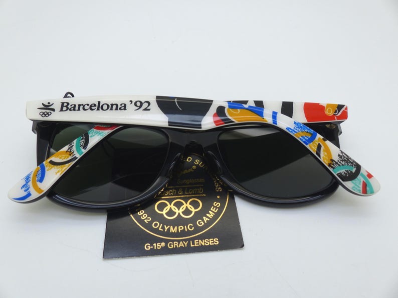 May include: Black sunglasses with dark lenses. The white arms are decorated with colorful abstract designs and the text "Barcelona '92". A tag with the Olympic rings and "992 Olympic Games" is visible.