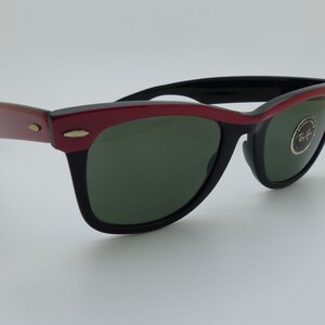 Puede incluir: Gafas de sol con montura negra y borde superior rojo. Las lentes son verdes y las patillas son de color marr&oacute;n rojizo. Las gafas tienen un dise&ntilde;o cl&aacute;sico y retro. El logotipo de Ray-Ban es visible en la lente.