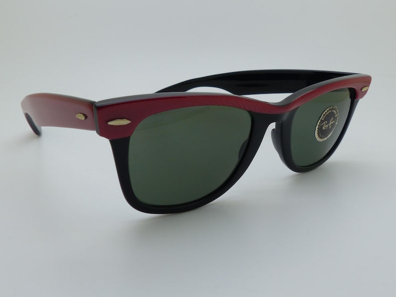 Puede incluir: Gafas de sol con montura negra y borde superior rojo. Las lentes son verdes y las patillas son de color marr&oacute;n rojizo. Las gafas tienen un dise&ntilde;o cl&aacute;sico y retro. El logotipo de Ray-Ban es visible en la lente.