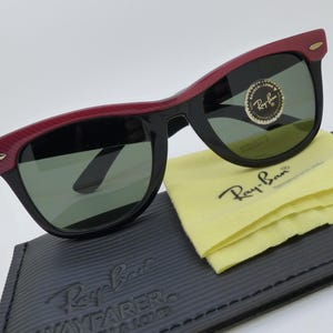 Puede incluir: Gafas de sol Ray-Ban Wayfarer negras con detalles rojos en la parte superior de la montura. Las gafas descansan sobre un estuche negro con el logotipo de Ray-Ban y un pa&ntilde;o de limpieza amarillo. Las lentes son verdes.