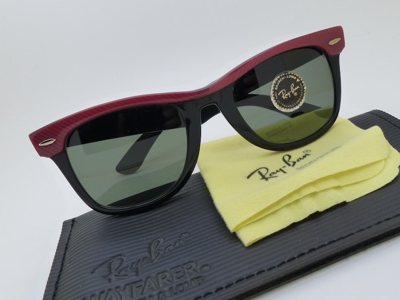 Puede incluir: Gafas de sol Ray-Ban Wayfarer negras con detalles rojos en la parte superior de la montura. Las gafas descansan sobre un estuche negro con el logotipo de Ray-Ban y un pa&ntilde;o de limpieza amarillo. Las lentes son verdes.