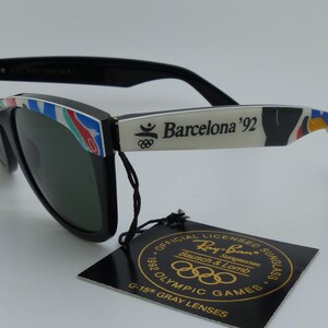 May include: Black Ray-Ban sunglasses with green lenses. The arms are white with "Barcelona '92" text and colorful accents. A tag reads "Official Licensed Sunglasses 1992 Olympic Games".