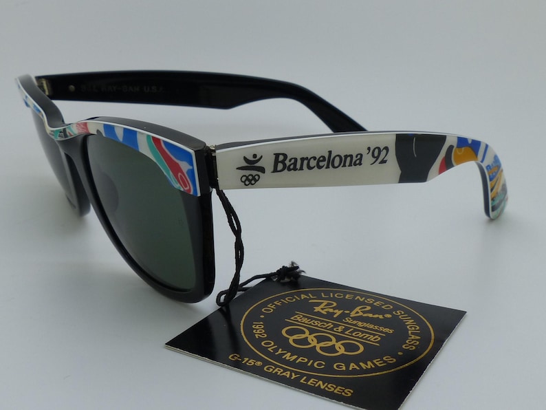 May include: Black Ray-Ban sunglasses with green lenses. The arms are white with "Barcelona '92" text and colorful accents. A tag reads "Official Licensed Sunglasses 1992 Olympic Games".