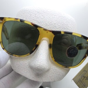 May include: Tortoiseshell-patterned sunglasses with green lenses. The sunglasses are displayed on a white mannequin head. A tag with the Ray-Ban logo is attached to the sunglasses.
