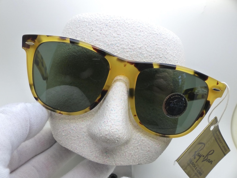 May include: Tortoiseshell-patterned sunglasses with green lenses. The sunglasses are displayed on a white mannequin head. A tag with the Ray-Ban logo is attached to the sunglasses.