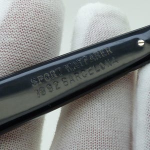May include: Close-up of a dark gray eyeglass arm with the text "SPORT WAYFARER 1992 BARCELONA" engraved on the surface. The arm is held against a white fabric background.