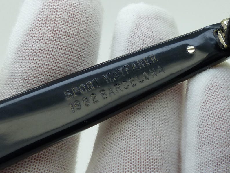 May include: Close-up of a dark gray eyeglass arm with the text "SPORT WAYFARER 1992 BARCELONA" engraved on the surface. The arm is held against a white fabric background.