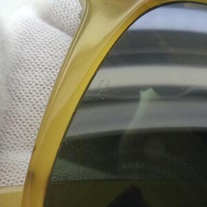 May include: Close-up of a pair of sunglasses with a yellow-tinted frame and dark lenses. The sunglasses are held against a white, textured background. The lenses have a slight reflection.