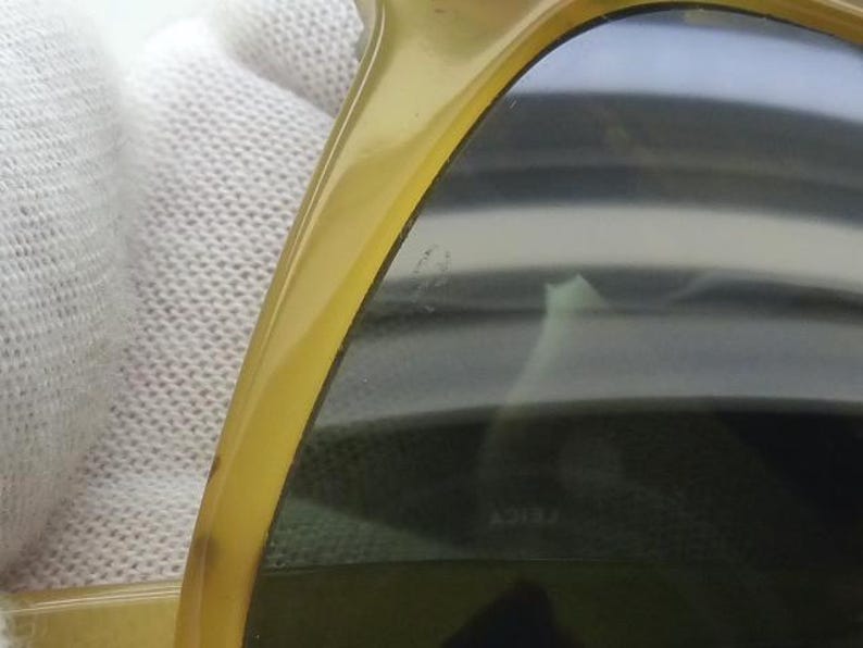 May include: Close-up of a pair of sunglasses with a yellow-tinted frame and dark lenses. The sunglasses are held against a white, textured background. The lenses have a slight reflection.