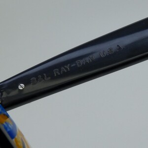 May include: Close-up of a black Ray-Ban sunglasses arm. The text "B&L RAY-BAN U.S.A." is engraved on the arm. The sunglasses arm is dark gray and has a shiny finish. The image is well-lit and the details are clear.