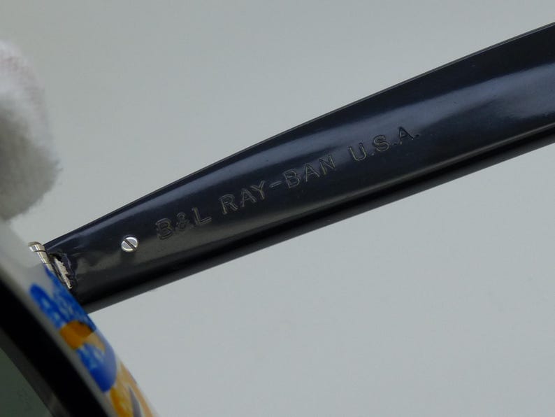 May include: Close-up of a black Ray-Ban sunglasses arm. The text "B&L RAY-BAN U.S.A." is engraved on the arm. The sunglasses arm is dark gray and has a shiny finish. The image is well-lit and the details are clear.