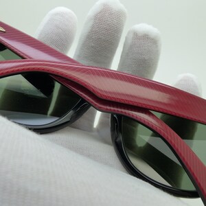 Puede incluir: Primer plano de gafas de sol con montura roja texturizada y bordes negros. Las lentes son de color verde oscuro. Las gafas de sol est&aacute;n sujetas por una mano enguantada.