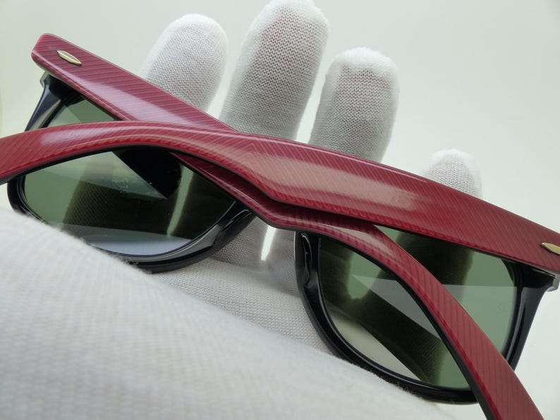 Puede incluir: Primer plano de gafas de sol con montura roja texturizada y bordes negros. Las lentes son de color verde oscuro. Las gafas de sol est&aacute;n sujetas por una mano enguantada.