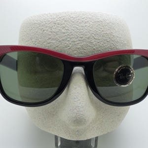 Puede incluir: Gafas de sol negras con montura superior roja y lentes verdes. Las gafas de sol tienen un dise&ntilde;o angular cl&aacute;sico. El logotipo de Ray-Ban es visible en la lente derecha. Las gafas se exhiben en una cabeza de maniqu&iacute;.