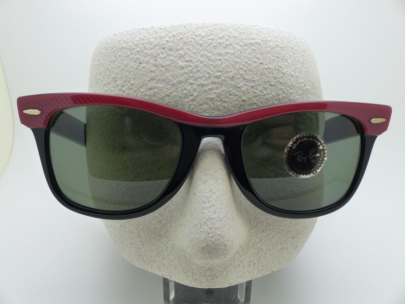 Puede incluir: Gafas de sol negras con montura superior roja y lentes verdes. Las gafas de sol tienen un dise&ntilde;o angular cl&aacute;sico. El logotipo de Ray-Ban es visible en la lente derecha. Las gafas se exhiben en una cabeza de maniqu&iacute;.