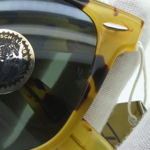 May include: Close-up of sunglasses with a tortoiseshell frame. The lenses are dark, and a gold sticker with text is visible on the lens. The frame has a silver accent and a tag attached.