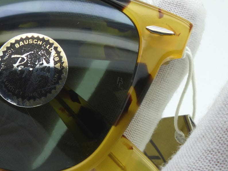 May include: Close-up of sunglasses with a tortoiseshell frame. The lenses are dark, and a gold sticker with text is visible on the lens. The frame has a silver accent and a tag attached.
