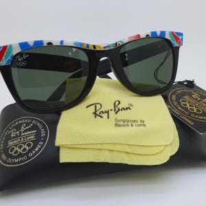May include: Black Ray-Ban sunglasses with a colorful patterned design on the frames. The sunglasses rest on a black case with a yellow cleaning cloth. The sunglasses are labeled as official licensed 1992 Olympic Games.