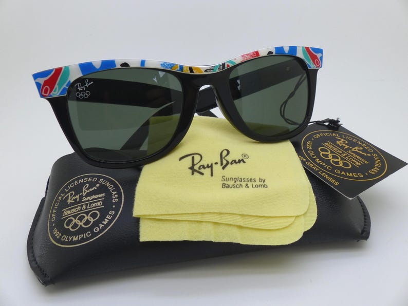 May include: Black Ray-Ban sunglasses with a colorful patterned design on the frames. The sunglasses rest on a black case with a yellow cleaning cloth. The sunglasses are labeled as official licensed 1992 Olympic Games.