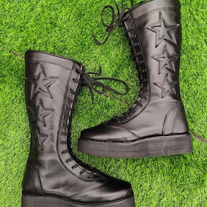 May include: A pair of black, knee-high boots with star-shaped cutouts. The boots have a lace-up design and thick soles. The boots are made of a smooth material and are set against a green background.