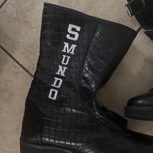 May include: Black, high-top boots with a textured, crocodile-like pattern. The boots have a zipper and the word "SMUNDO" in white block letters on the side. The boots are on a tiled floor.