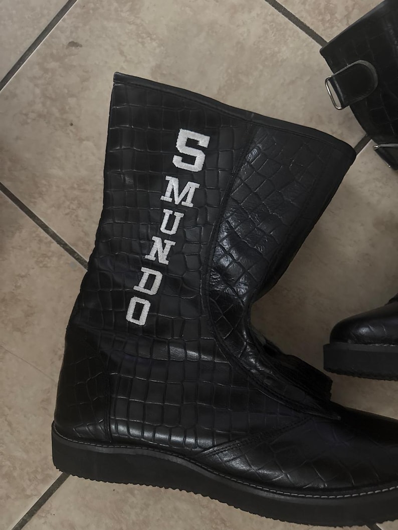 May include: Black, high-top boots with a textured, crocodile-like pattern. The boots have a zipper and the word "SMUNDO" in white block letters on the side. The boots are on a tiled floor.