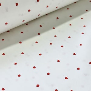 May include: White fabric featuring a repeating pattern of small red hearts. The material is rolled up in the upper left, showing more of the heart-printed fabric. The hearts are evenly spaced across the white background.