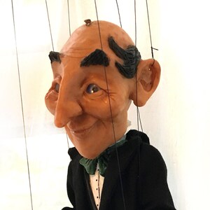 May include: A hand-crafted puppet of a man wearing a black suit with a green bow tie. The puppet has a large nose and a friendly expression.