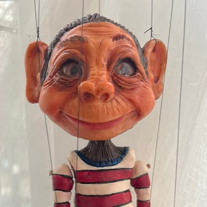 May include: A handcrafted marionette puppet with a detailed, expressive face and a striped shirt. The puppet's skin is a warm, reddish-brown, and it has articulated limbs and hands. The puppet is suspended by strings.
