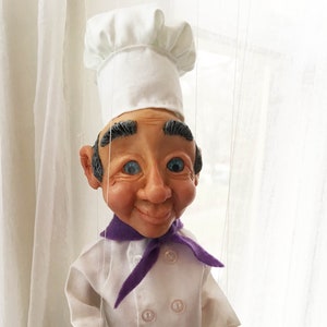 May include: A white chef puppet with a purple scarf, black and white checkered pants, and black shoes. The puppet is holding a small metal whisk in its right hand.