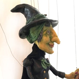 Puede incluir: Un títere de bruja verde y negro con un sombrero puntiagudo, sosteniendo un bastón de madera. El títere tiene una cara verde con una nariz larga y lleva un vestido negro con un lazo verde.