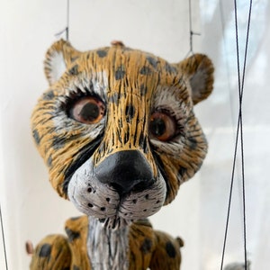 May include: A hand-crafted puppet of a cheetah with a spotted fur pattern. The puppet has large, expressive eyes and a detailed face. The puppet is made of a textured material and is suspended by strings.