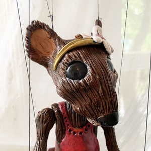 May include: A brown puppet mouse wearing a red dress with a gold trim and a gold headband with a white flower. The mouse has large black eyes and a brown nose.