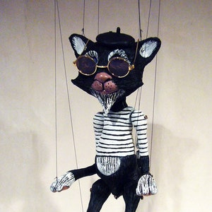 May include: A black and white puppet cat wearing a black beret and sunglasses. The cat is wearing a striped shirt and has a white beard. The puppet is suspended by strings.