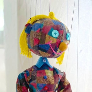 Może przedstawiać: Kolorowa kukiełka z patchworku z żółtymi włosami, niebieskimi oczami i czerwonym noskiem z guzika. Kukiełka ma na sobie wielokolorową sukienkę i czerwone buty.