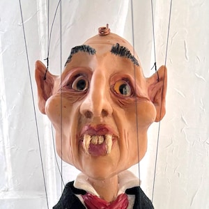 May include: A handcrafted marionette puppet of a vampire, featuring a detailed face with fangs, large ears, and a bald head. The puppet wears a black suit, white shirt, and a red ascot. The puppet's hands and feet are also detailed.