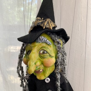 Puede incluir: Títere de bruja hecho a mano con cara verde, pelo gris y sombrero negro adornado con un murciélago dorado. La bruja lleva un vestido negro con tres botones. El títere está suspendido por hilos negros.
