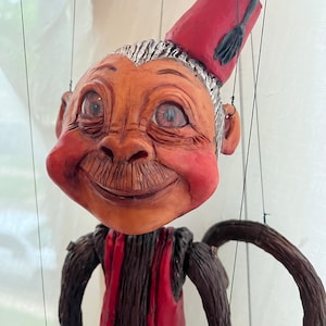 May include: A hand-crafted puppet with a brown wooden body, a red waistcoat, and a red fez with a black tassel. The puppet has a smiling face with brown eyes, a moustache, and rosy cheeks.